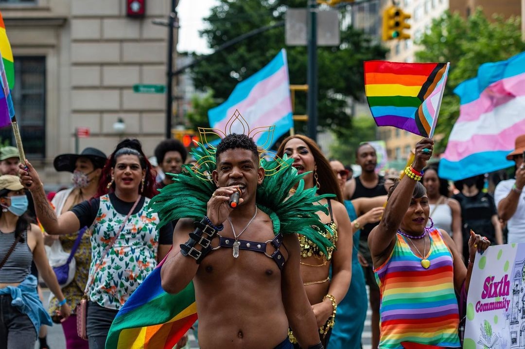 Rohan Zhou-Lee leads a group of colorfully and playfully dressed people in a parade. Hand on mic proudly speaking